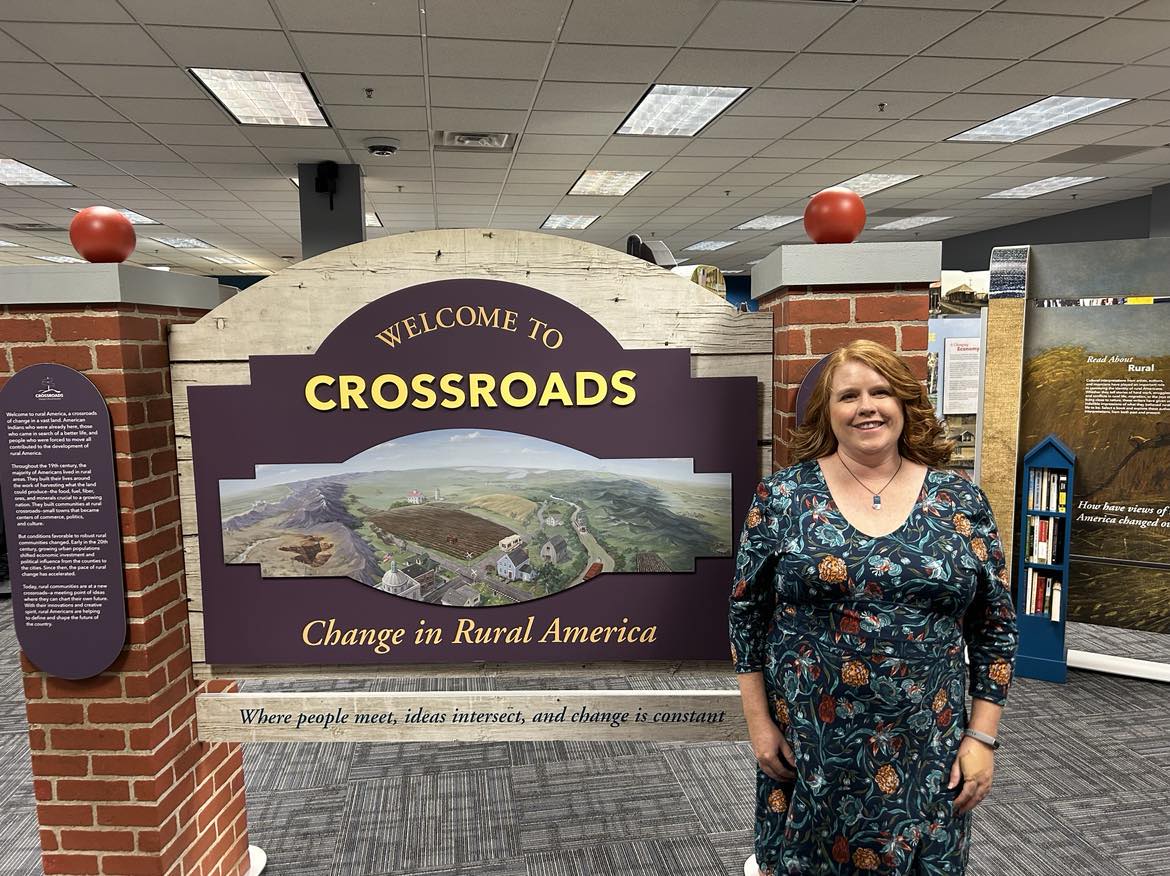 woman in a fower dress standing in front of a big sign for the crossroads exhibit
