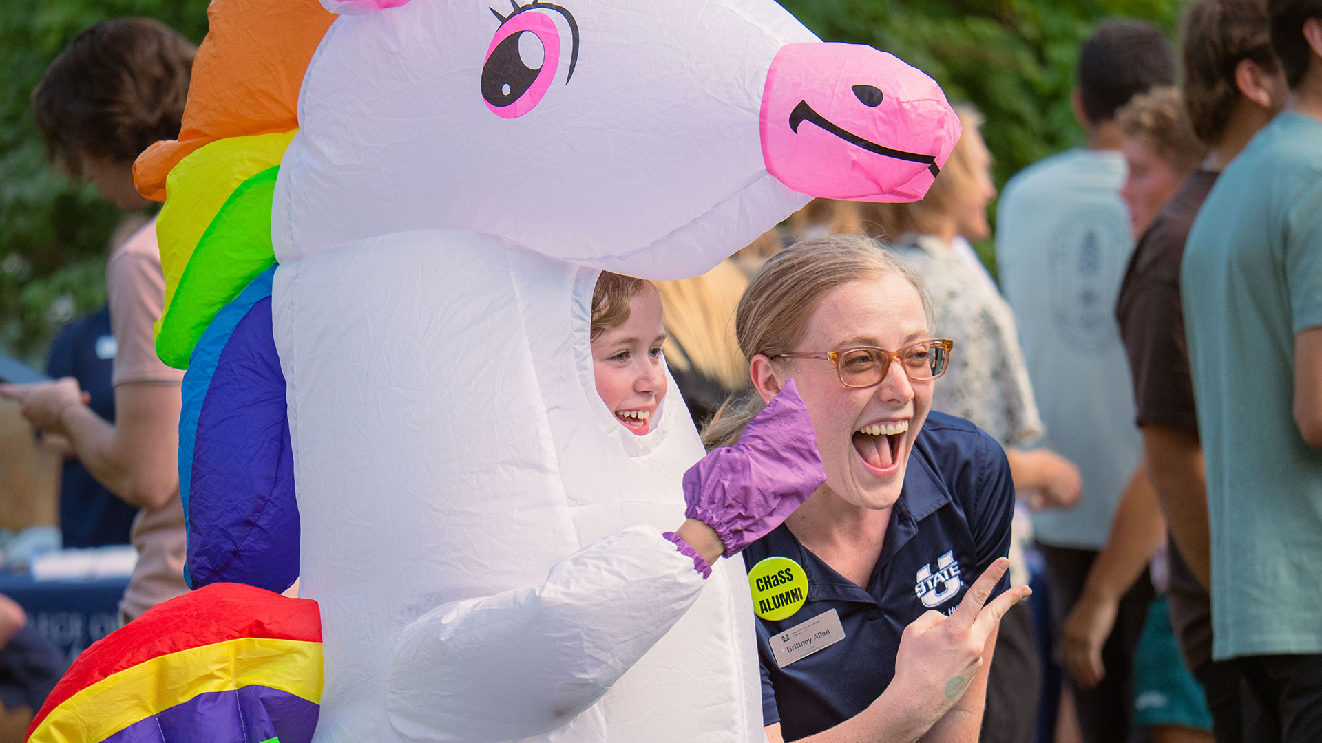 A little girl in a inflatable unicorn costume and a chass alumni pose smiling brightly for a picture in front of crowds of students visiting booths