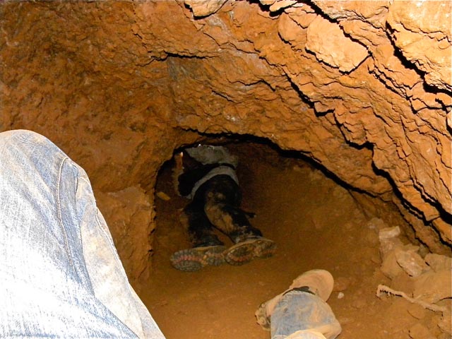 A man lying in a dirt tunnel mining gold. In the foreground there are the legs of another man who appears to be taking the photo.