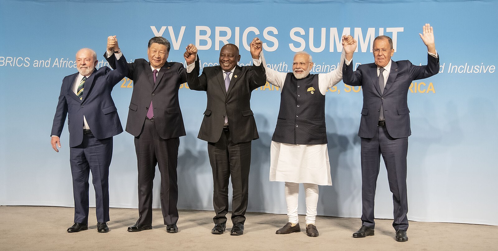 Indian, Brazilian, Chinese, Russian, and South African leaders standing in front of a row of flags with hands raised together in the air at the BRICS Summit 2023.