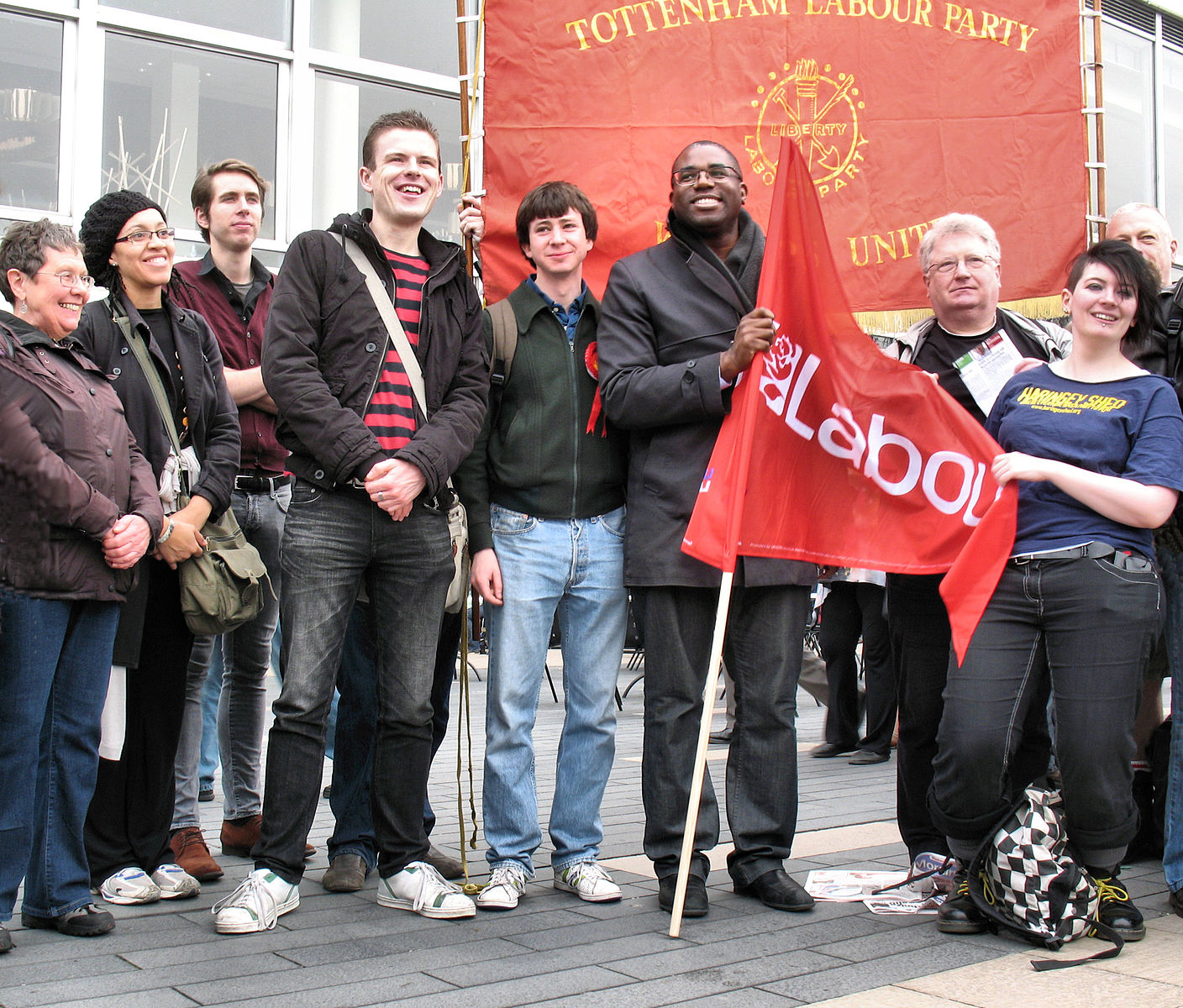 David Lammy standing in the center of a line of Labour Party supporters in London in front of a red brick wall and the Labour Party flag. 