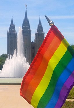 Gay pride flag focused in front of the SLC Temple and a fountain on a sunny day