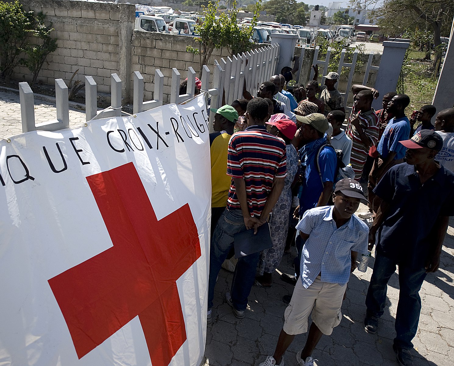 A group of Haitian earthquake survivors waiting outside a white picket near a red cross station in Haiti for food.