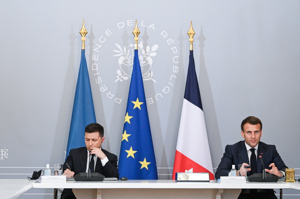 Emmanuel Macron and Volodymyr Zelenskyy sitting at a table during a press conference in front of a grey backdrop.
