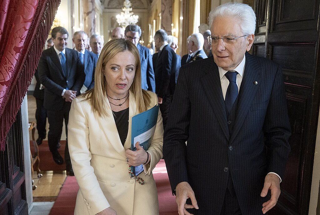 Italian Prime Minister Giorgia Meloni walking with Italian President Sergio Mattarella through dark brown doors with Italian members of government in the background. 