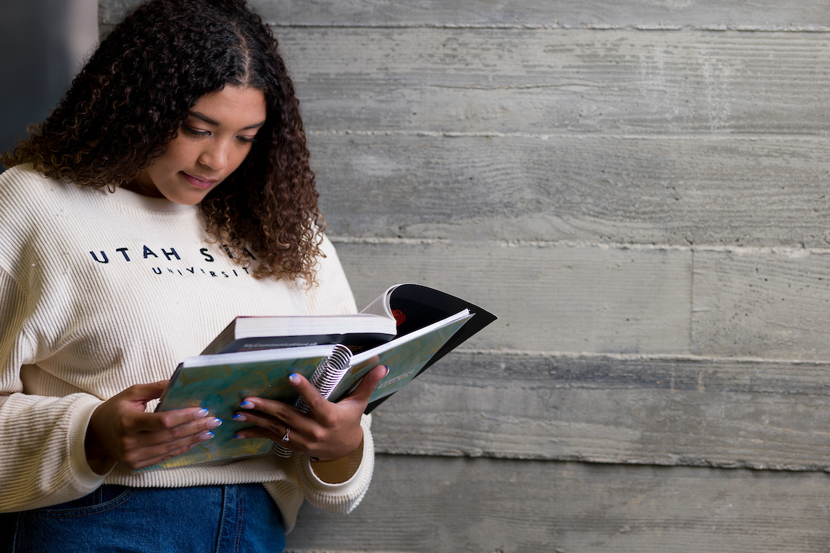 A USU student looks down at a book, as she reads while leaning against a wall.